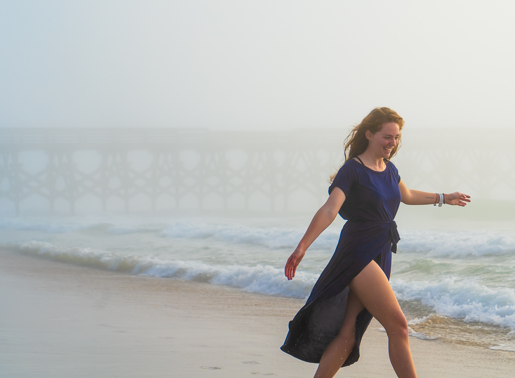 Woman candidly walking along the shore of a beach in a blue dress with a pier in the background