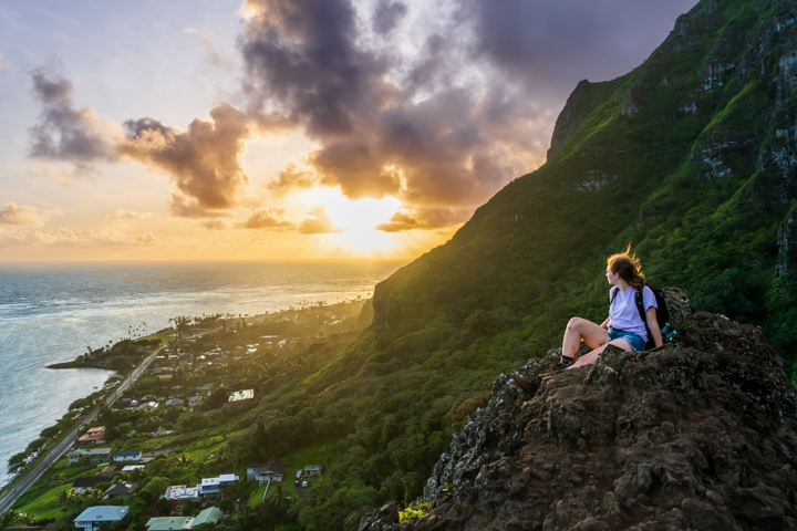 Woman sitting on a rock formation overlooking hawaiian mountains and the pacific ocean at sunrise