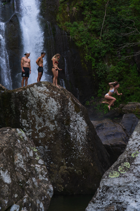Girl jumping off of a rock in front of a waterfall with her friends queued up to jump next