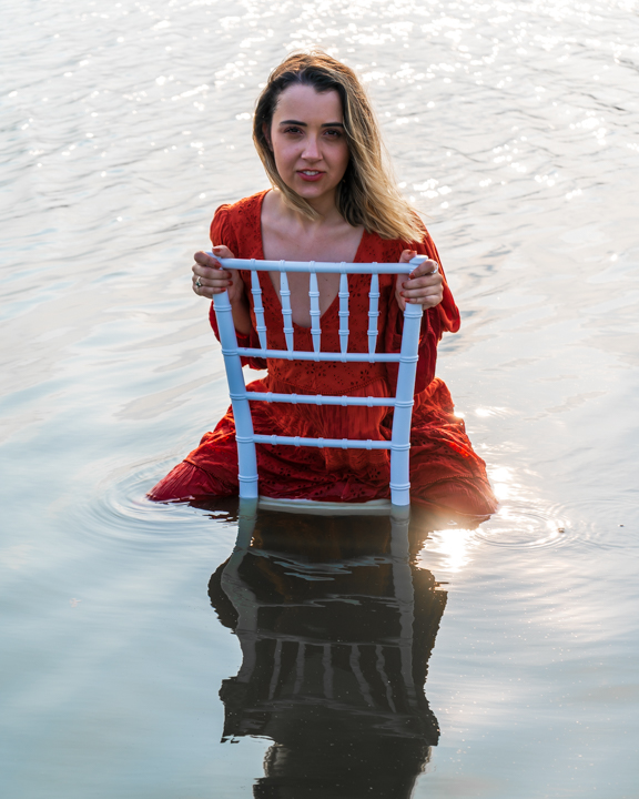 Woman siting on a backwards chair half submerged in a lake, she is wearing an orange dress