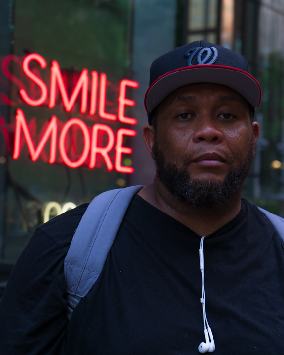 Man with a hat looking strait faced in front of a neon sign saying smile more