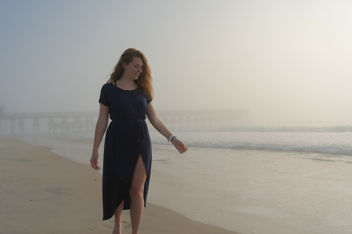 Woman walking on a foggy beach in front of a pier looking down her outstretched arm into the water